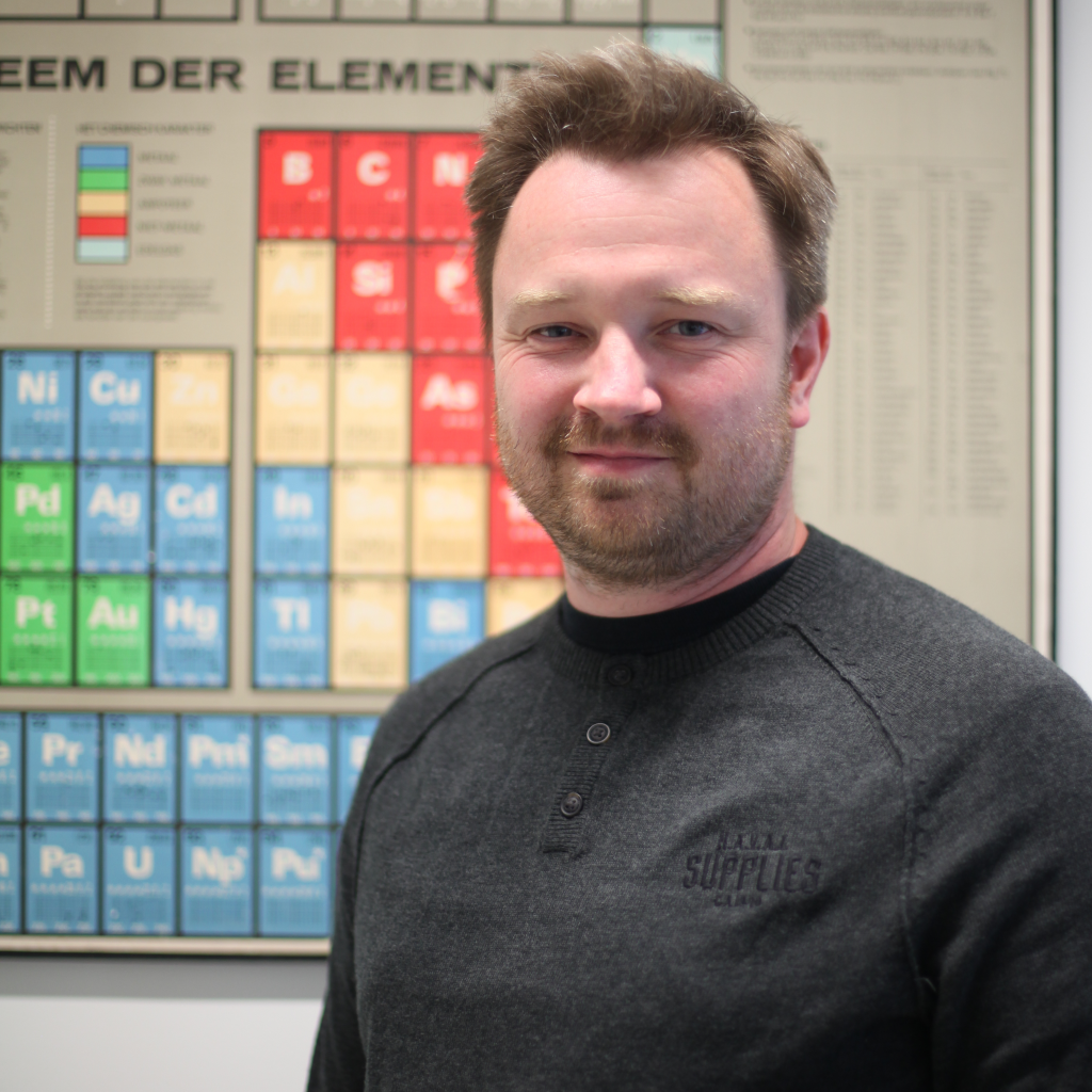 softly smiling man with short brown hair and beard in front of a periodic table
